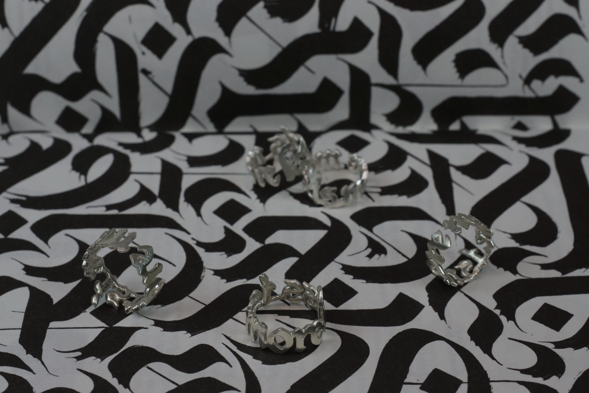 Five Sterling 925 silver rings engraved with 'more is more,' and 'less is more,' arranged on a black-and-white background featuring calligraphy in blackletter font. The intricate shape of the rings contrast beautifully with the bold, dynamic strokes of the blackletter design.