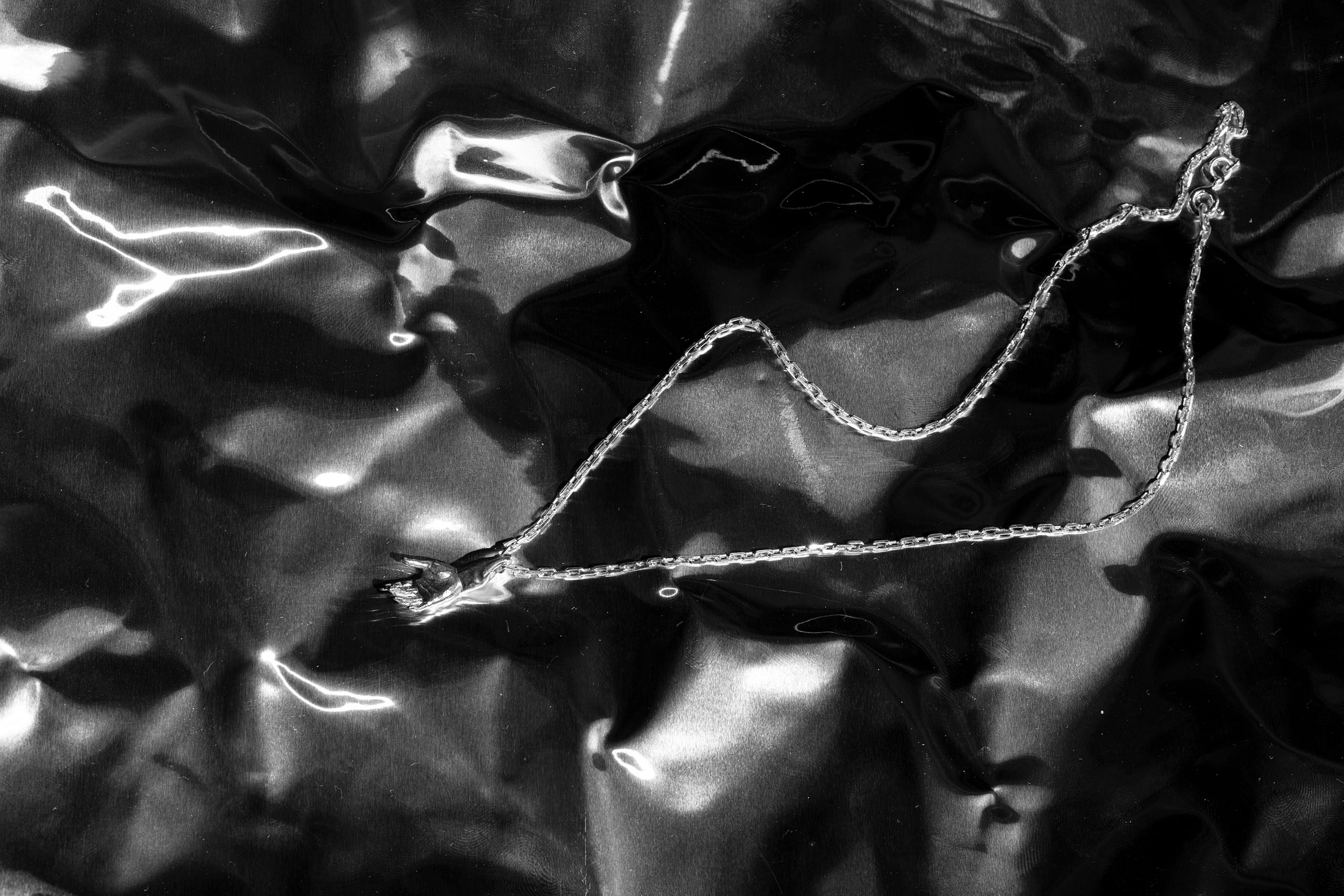 Sterling 925 silver pendant of 'The Hand of Adam,' hanging from a silver chain, placed on a shiny, crumpled black background. The detailed design of the hand is prominently displayed, with reflections from the glossy surface adding texture and depth to the composition.