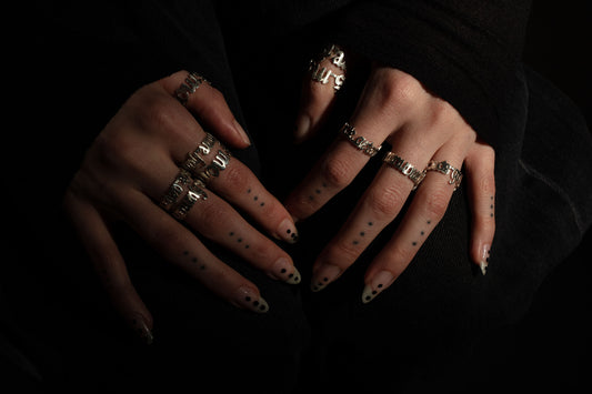 Sterling 925 silver rings, engraved with 'more is more,' 'less is more,' 'sub specie aeternitatis,' and 'errare humanum est,' worn on fingers with dotted tattoos and stylish nails, set against a dark background.