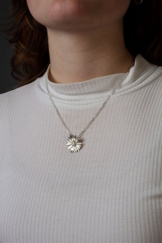 Sterling 925 silver pendant of a daisy flower, hanging from a delicate silver chain around the neck. The pendant's intricate floral design is clearly visible, with the person wearing a white ribbed shirt, highlighting the contrast between the bright pendant and the soft texture of the fabric.
