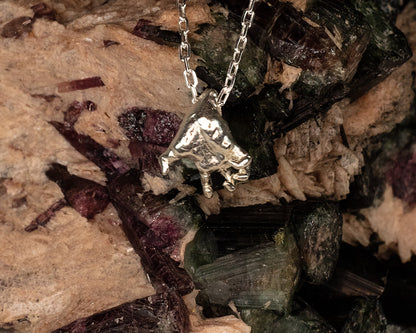 Sterling 925 silver pendant of Rodin's sculpture 'Clenched Hand,' hanging from a chain, set against a textured backdrop of colorful mineral crystals.