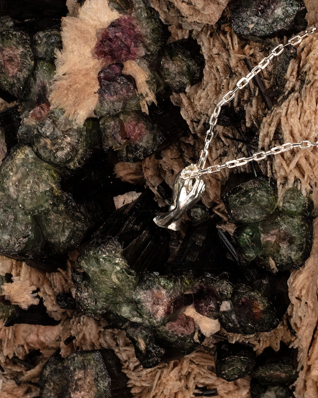 Sterling 925 silver pendant of Rodin's sculpture 'The Cathedral,' hanging from a chain and placed on a textured backdrop of mineral crystals with green and black hues.