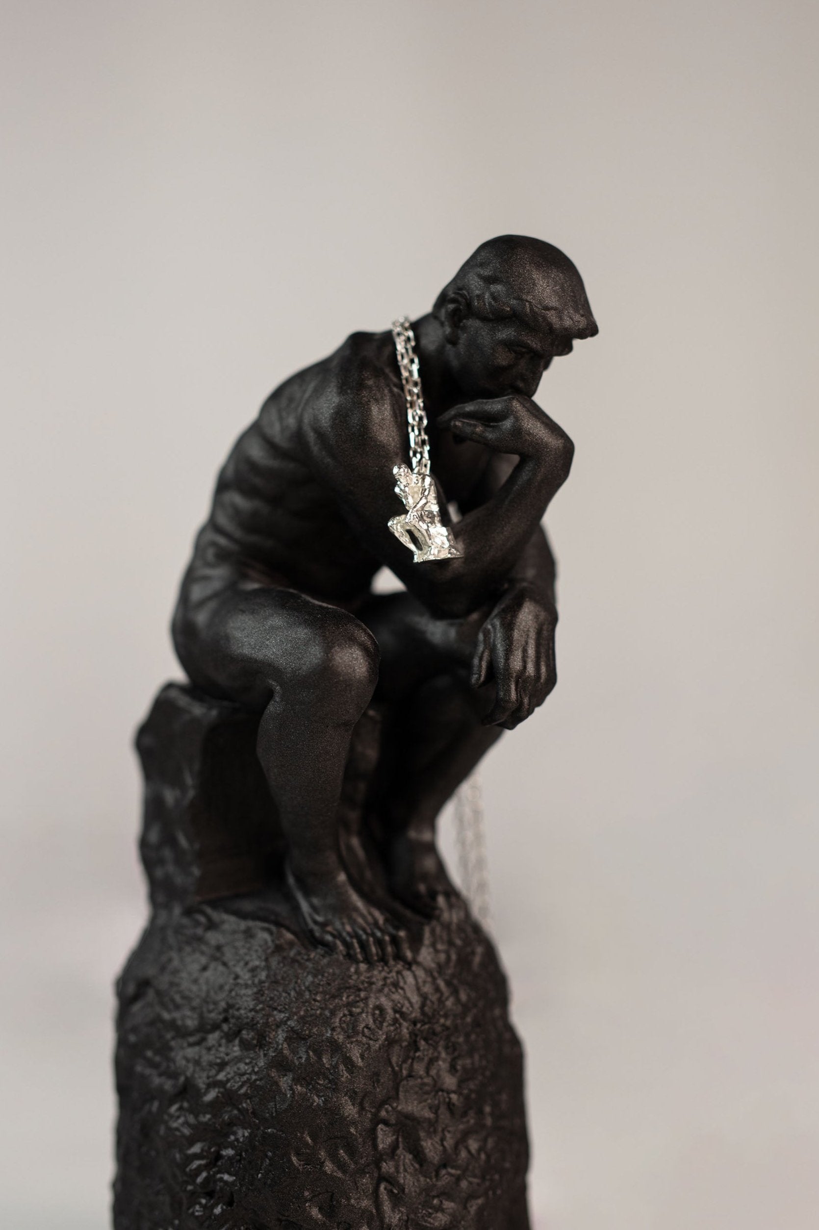 Small 925 sterling silver pendant of 'The Thinker' statue by Rodin hanging from a chain around the neck of a black sculpture of the same figure, set against a neutral background.