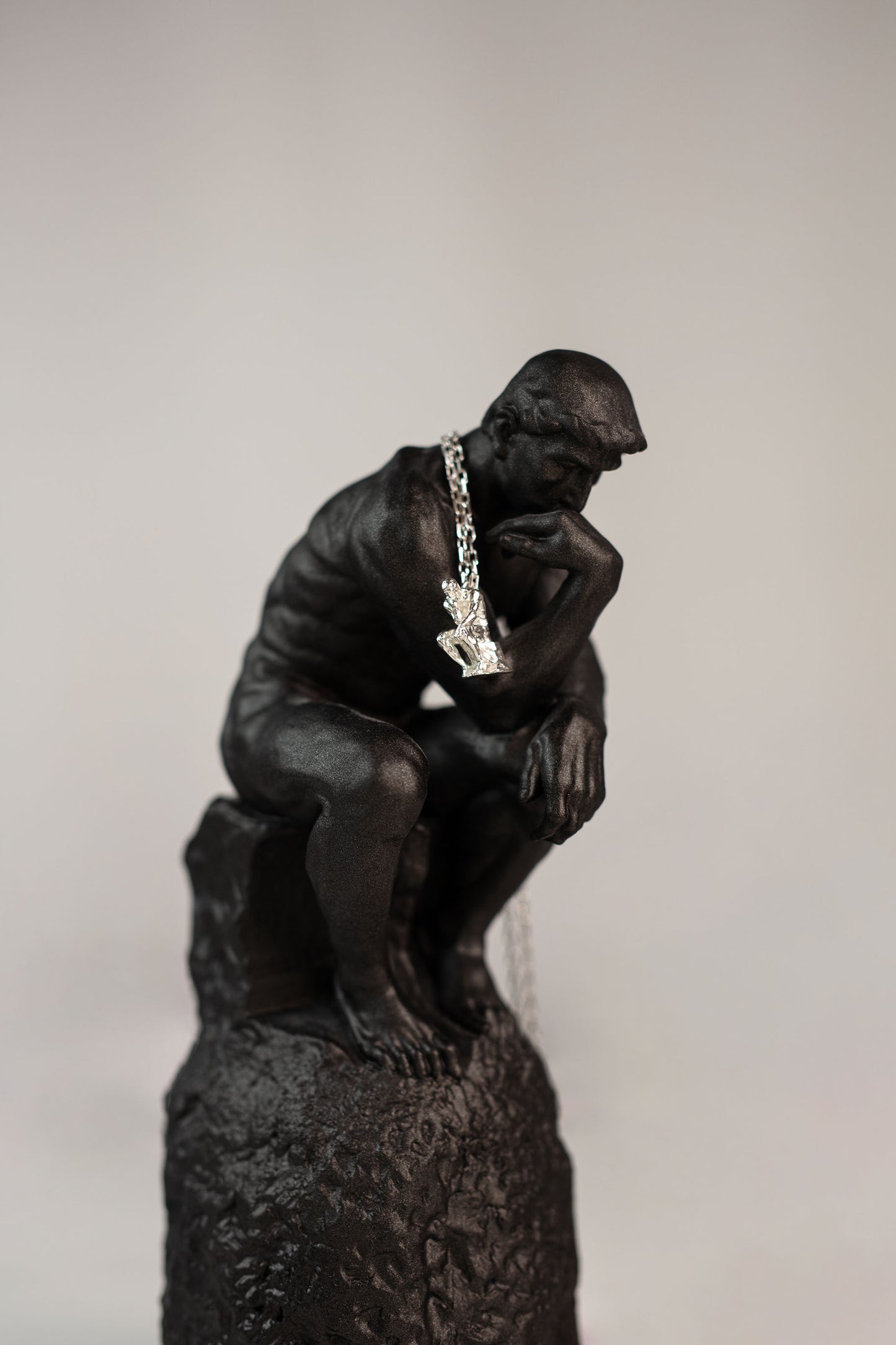 Small 925 sterling silver pendant of 'The Thinker' statue by Rodin hanging from a chain around the neck of a black sculpture of the same figure, set against a neutral background.