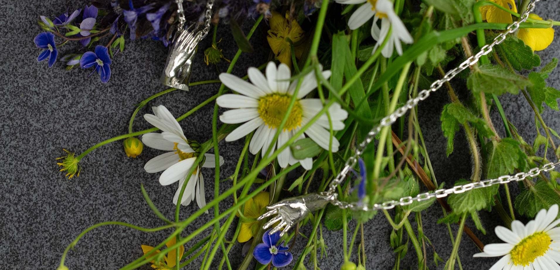 Sterling 925 silver pendants of Rodin's sculpture 'The Cathedral' and 'The Hand of Adam' displayed among wildflowers, including vibrant yellow buttercups, delicate purple violets, soft blue forget-me-nots, and white daisies. The intricate designs of the hands, symbolizing connection and creation, are showcased against the natural beauty of the flowers and a dark gray background.