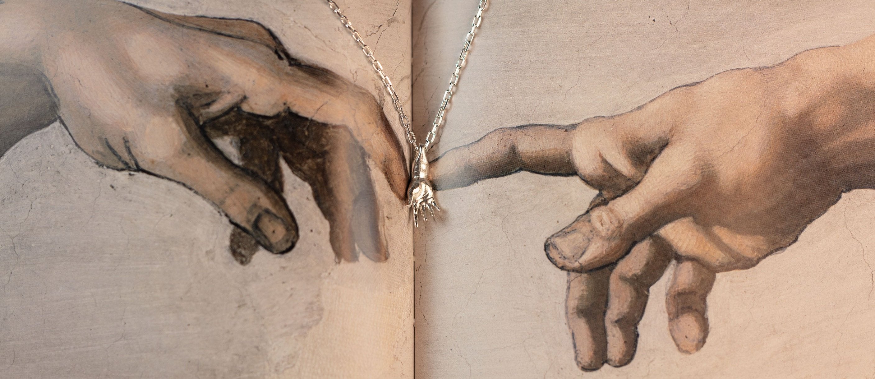 Close-up of two hands from Michelangelo's 'The Creation of Adam,' with a sterling silver necklace featuring a hand-shaped pendant placed between the fingers.