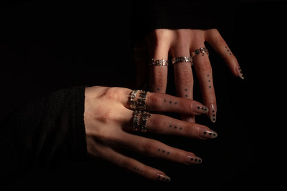 Two hands adorned with multiple Sterling 925 silver rings, engraved with the phrases 'more is more,' 'less is more,' 'sub specie aeternitatis,' and 'errare humanum est.' The rings are worn on fingers with dotted tattoos and stylish nails, set against a dark background that highlights the intricate designs of the rings.