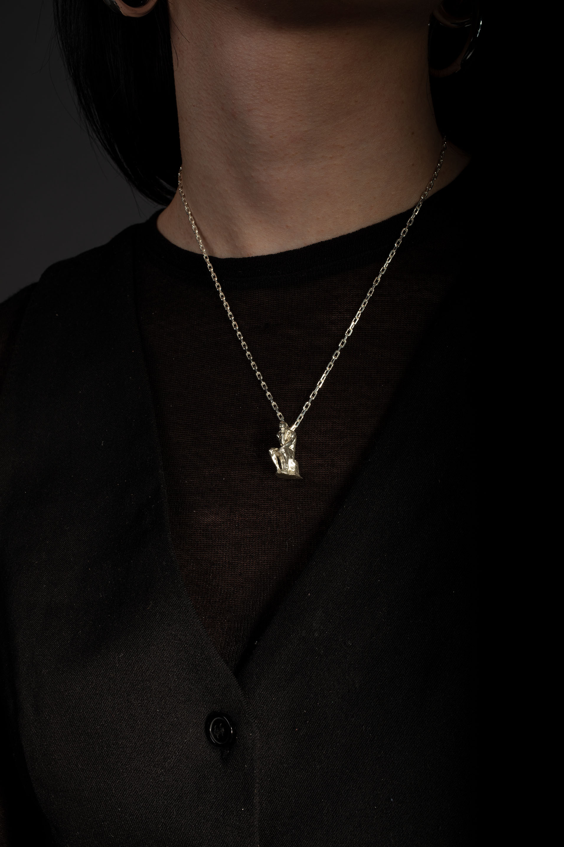 Sterling 925 silver pendant modeled after Rodin's sculpture 'The Thinker,' hanging from a delicate chain around the neck of the wearer, who is dressed in a black top. The pendant, depicting the seated figure in a contemplative pose, stands out against the dark background.