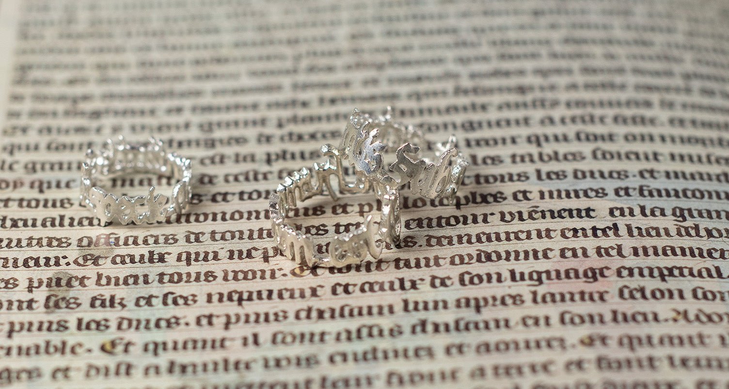 Two 925 sterling silver rings with engraved Latin phrases: one with 'sub specie aeternitatis' and the other with 'errare humanum est,' placed on an old manuscript with faded, intricate text in the background.