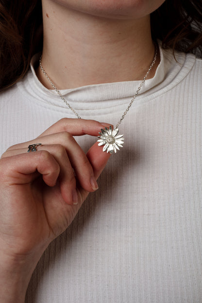 Sterling 925 silver pendant of a daisy flower, held in hand and hanging from a delicate silver chain. The pendant's intricate floral design is clearly visible, with the person wearing a white ribbed shirt, highlighting the contrast between the bright pendant and the soft texture of the fabric.